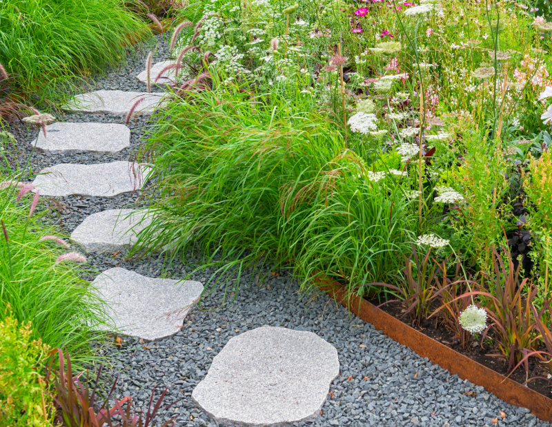 Ein Gartenweg aus Trittsteinen in Kiesbett, an dessen Seiten sich eingefasste Staudenbeete befinden.